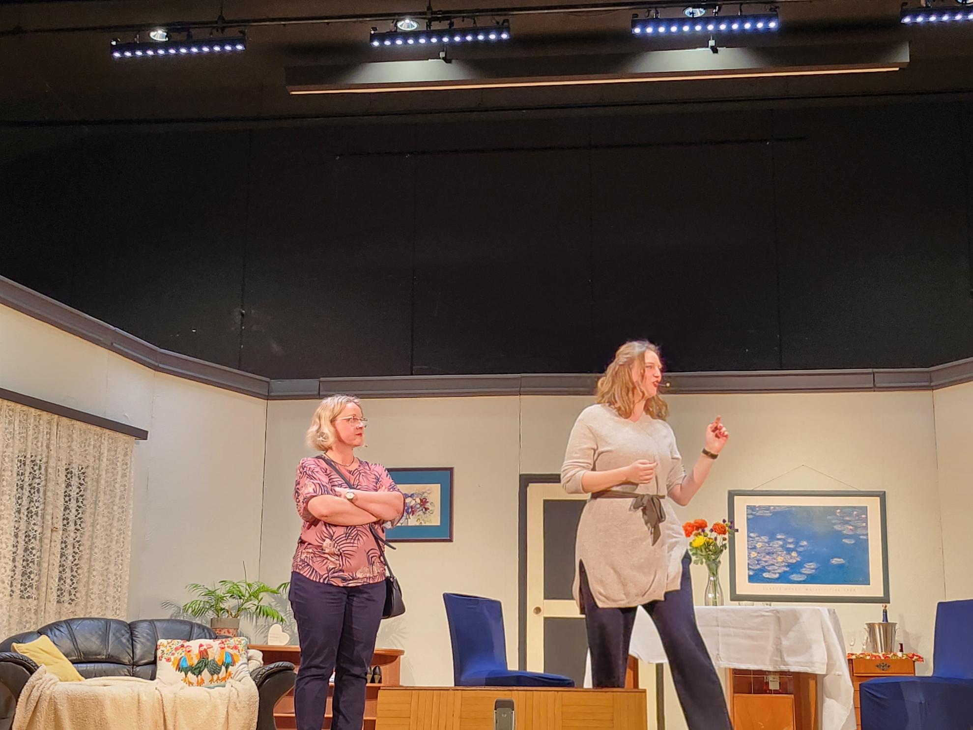 2 Frauen stehen vor einem festlich gedeckten Tisch, die Rechte wendet der Linken den Rücken zu, im Hintergrund ist die Theaterkulisse zu sehen
