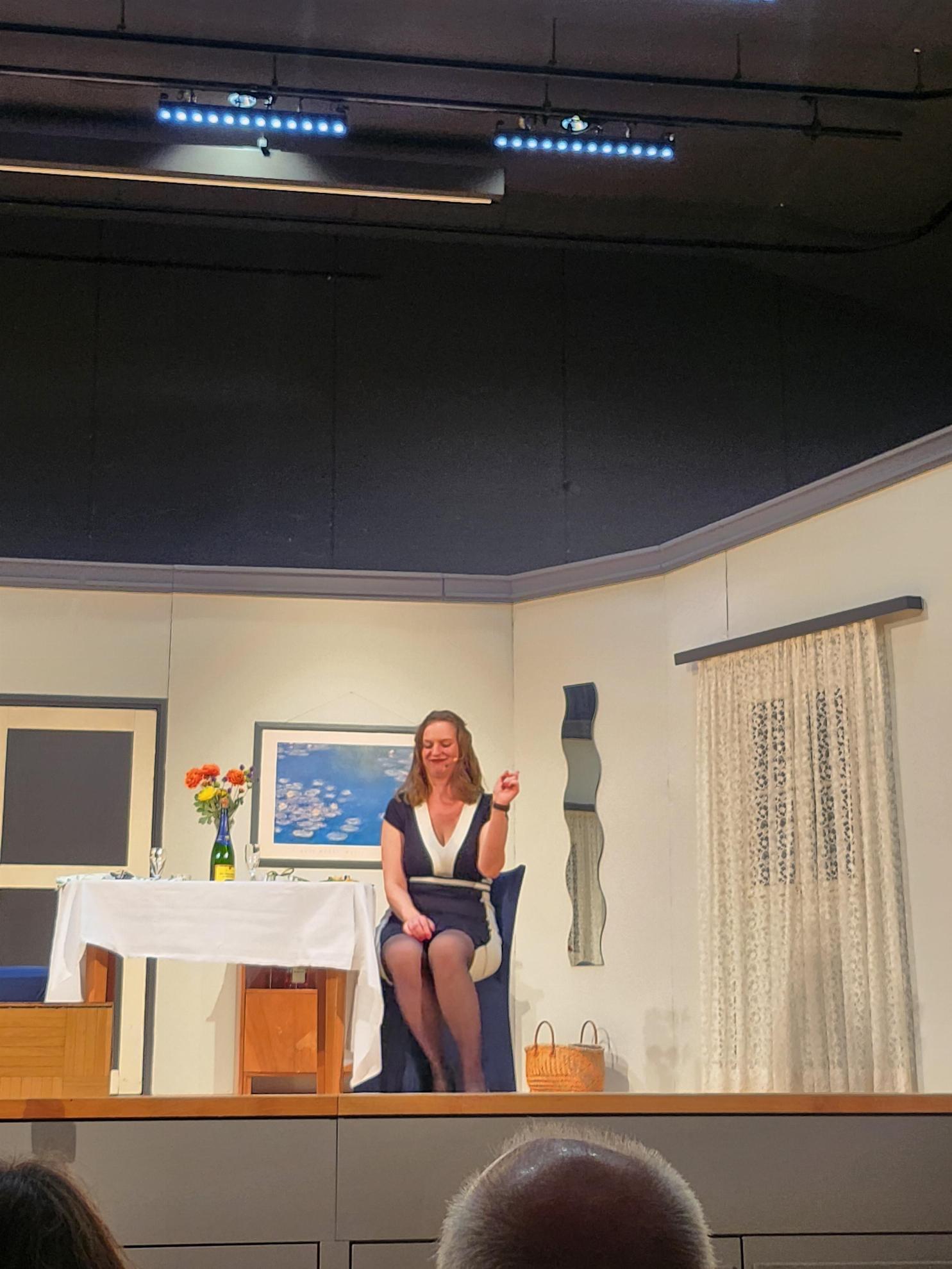 Eine schön gekleidete Frau sitzt an einem festlich gedeckten Tisch auf der Bühne, im Hintergrund die Theaterkulisse die die Innenansicht eines Zimmers darstellt