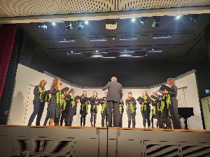 Der Frauenchor steht auf der Bühne, im Vordergrund steht der Dirigent mit dem Rücken zum Publikum, Rechts im Hintergrund steht ein Flügel