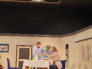 Ein stehender Mann und eine sitzende Frau an einem gedeckten Tisch auf der Bühne, im Hintergrund die Theaterkulisse die ein Zimmer darstellt