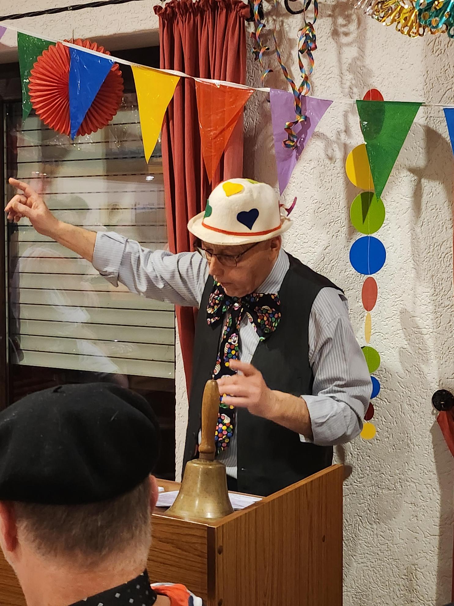 Der Moderator des Abends steht am Rednerpult auf dem eine Handglocke steht, über ihm bunte Wimpel