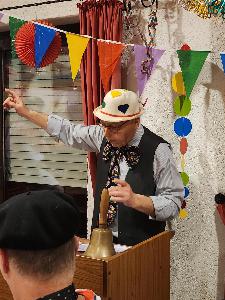 Der Moderator des Abends steht am Rednerpult auf dem eine Handglocke steht, über ihm bunte Wimpel