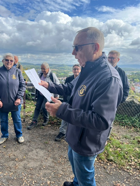Jemand steht im Vordergund, ein Notenblatt in der Hand und singt unter blauem Himmel mit weißen Wolken. Im Hintergrund stehen Leute.