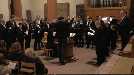 Ein Männerchor singt in der evangelischen Kirche in Schönau. Der Dirigent steht neben einem Klavier und hat die Arme zum Takt erhoben, die Pianistin begleitet den Chor.